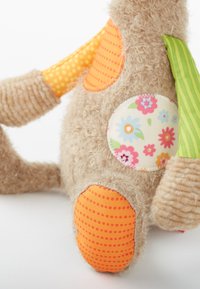 Jouet en peluche doux de couleur beige avec une fourrure texturée. Comprend des patches en tissu colorés en orange et vert, des motifs floraux et des pois.