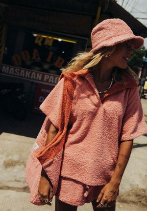 Różowy top z frotté z kołnierzykiem, w komplecie z pasującymi szortami i pomarańczową torbą. Charakteryzuje się luźnym krojem i fakturowaną powierzchnią.