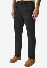 Pantalons plissés noirs en coton avec une coupe slim. Présente une fermeture à bouton et des poches latérales, associés à des chaussures en daim marron.