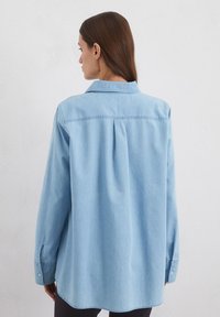 Lichtblauwe, lange mouwen spijkerblouse met een standaard kraag, achter plooi detail en knoopmanchetten, met een relaxte pasvorm.