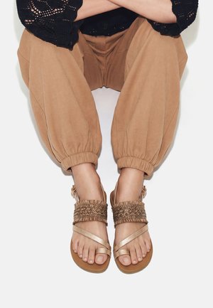Pies con sandalias beige con tiras trenzadas, combinadas con pantalones beige sueltos con puños elásticos y un top negro de crochet, piernas cruzadas.