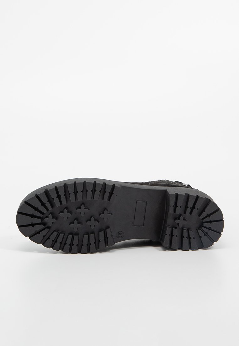 Suola di stivale in gomma nera rivolta verso l'alto, che mostra scanalature profonde con motivi rettangolari e a croce su uno sfondo bianco.