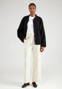 Zwarte oversized jas met drukknoopjes, gedragen over een witte blouse. Gecombineerd met crèmekleurige broeken met een strik taille en zwarte veterschoenen.