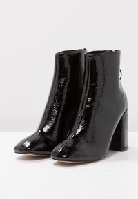 Schwarze Knöchelstiefel aus glänzendem Lackleder, mit spitzem Zehenbereich, hohem Blockabsatz und subtiler Textur. Reißverschluss auf der Rückseite.