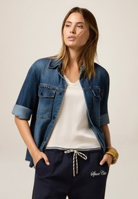 Denim jacket with rolled sleeves, studded shoulder accents. White V-neck shirt underneath, paired with navy drawstring pants featuring embroidery.