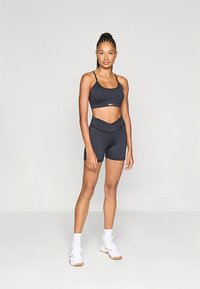 Zwarte sportbeha met dunne bandjes en bijpassende high-waisted shorts, beide van een gladde stof. Model draagt witte sportschoenen en sokken.