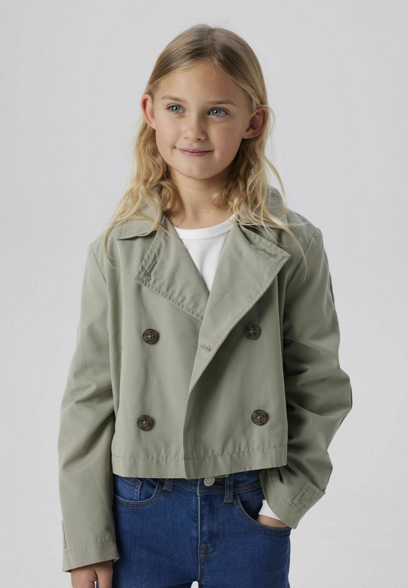 Chaqueta corta de doble botonadura en tejido verde claro con dos grandes botones, con solapas anchas y mangas largas. Llevada sobre una blusa blanca.