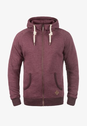 Sudadera con cremallera de color marrón con cordones en crema, bolsillos laterales y ribete marrón. Fabricada con una tela texturizada que presenta un pequeño logo en la parte inferior frontal.