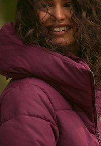 Bordo puffa jakna s visokim ovratnikom i zatvaračem, s kvadratnim uzorkom i mekim materijalom, vidljiva oko vrata i ramena.