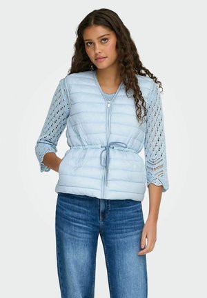 Kvinde iført lyseblå quiltet vest over blondeløs langærmet top og blå jeans, stående med den ene hånd i lommen mod en hvid baggrund.