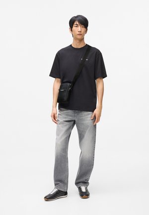 Ung mand stående iført sort T-shirt, grå falmede jeans, sorte sneakers og en sort crossbody-taske mod hvid baggrund.