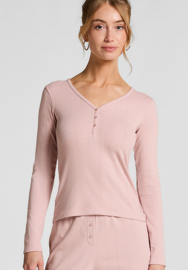 Jonge vrouw met lichtrood haar die een lichtroze, lange mouwen gebreide top en bijpassende onderkleding draagt, voor een effen lichtgrijze achtergrond.