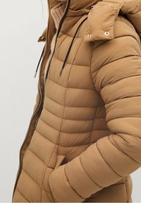 Tan pufferjakke med kviltet design, der har hætte, lynlås og lommer. Glat tekstur med slim-fit form og gyldne detaljer.