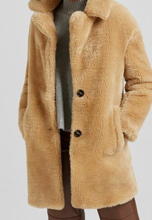 Tan faux fur jas met een zachte textuur, met een notch-kraag, twee knoppen aan de voorkant en zijzakken. Draagt over een grijze trui.