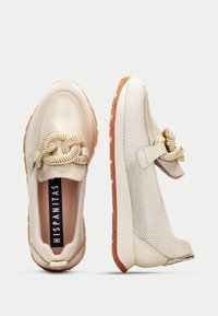 Beige mesh slip-on skor med en krämfärgad lädertå. Har en dekorativ flätad knut och en strukturerad brun gummisula. Varumärkeslogotyp synlig.