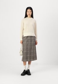 Maglione a maglia testurizzata di colore crema con scollo rotondo, abbinato a una gonna midi a quadri marrone. Scarpe nere e una piccola borsa bianca completano il look.