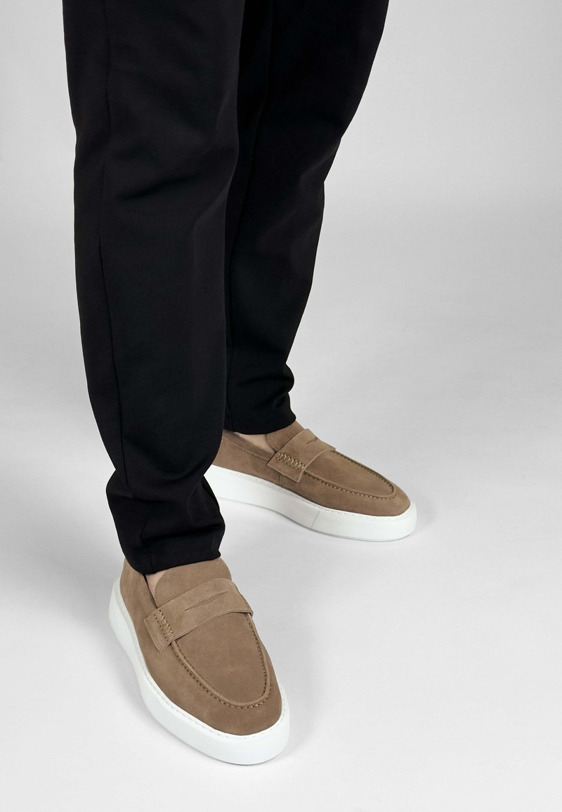Bruine suède instapschoenen met witte rubberen zolen, voorzien van een verhoogde naaddetail en een eenvoudig, strak ontwerp, gecombineerd met zwarte broeken.