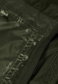Tessuto in nylon verde oliva con finitura lucida, caratterizzato da cuciture a punto, una cerniera nera e dettagli in velcro. È visibile un dettaglio della superficie texturizzata.
