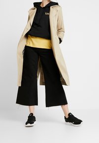 Bežový trenčkot přes černou mikinu a žluté tričko, doplněný černými širokými džíny a černými teniskami s bílou podrážkou.