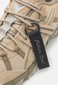 Beige trailová bota vyrobená z materiálů síťoviny a kůže, ploché béžové tkaničky, šedé akcenty a černý štítek s nápisem "Future Trail." Texturovaná podrážka.