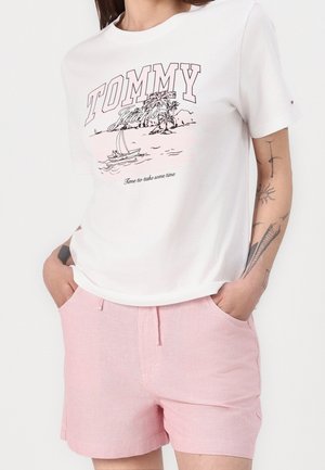Kobieta w białej koszulce z "Tommy Jeans" i motywem żaglówki oraz w jasnoróżowych szortach z rękami w kieszeniach.