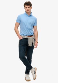 Lichtblauw polo-shirt met een opstaande kraag, gecombineerd met donkere jeans en witte sneakers. Een grijze trui is om de taille geknoopt.