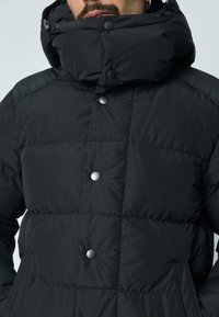 Doudoune noire avec col montant, boutons-pression, motif matelassé, texture lisse et mate, avec poches latérales.