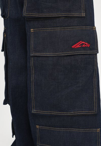 Tumed sinised denim cargo püksid mitme kasuliku taskuga ja vastandavate kollaste õmblustega. Ülemisel taskul on punane logo.