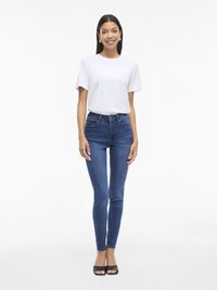 Camiseta blanca de manga corta combinada con unos jeans skinny oscuros ajustados. La modelo lleva sandalias negras tipo slide y pendientes de aro plateados.