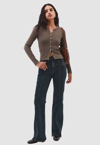 Brun långärmad knäpptröja i texturerat tyg, kombinerad med utsvängda mörka denimjeans med kontrasterande sömmar. Brun axelväska.