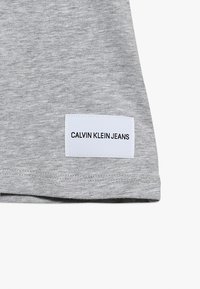 Šedá bavlněná látka s hladkou texturou, opatřená bílou etiketu s nápisem "CALVIN KLEIN JEANS" v černém písmu.