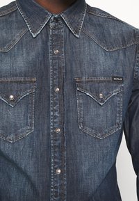 Granatowa koszula dżinsowa z pointed kołnierzykiem, zatrzaskami, podwójnymi kieszeniami na piersi oraz kontrastowymi detalami w szwie. Tkanina ma lekko chropowatą fakturę.