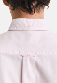 Ljusrosa skjorta med button-down krage, som har en bakfåll och en enkel knappstängning vid halsen. Slät tygtextur.