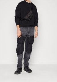 Sudadera negra con puños acanalados, pantalones cargo grises con múltiples bolsillos y botas negras gruesas, complementados por una riñonera negra.