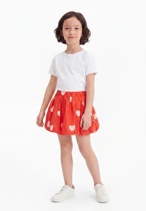 Enfant debout, mains détendues, portant un t-shirt blanc, une jupe rouge avec des cœurs blancs et des baskets blanches sur un fond uni.