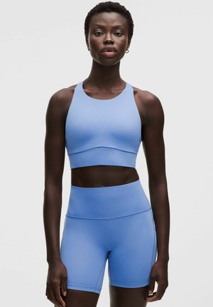 Vrouw draagt lichtblauwe sportbeha en hoge taille fietsbroek, staand tegen een effen grijze achtergrond.