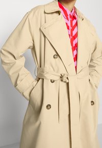 Beige double-breasted trench coat with belt tied at waist worn over a red and pink striped button-up shirt.