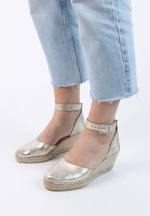 Metallic gold wedge espadrilles with ankle straps, featuring a closed toe and woven jute midsole. Worn with light blue cropped jeans.