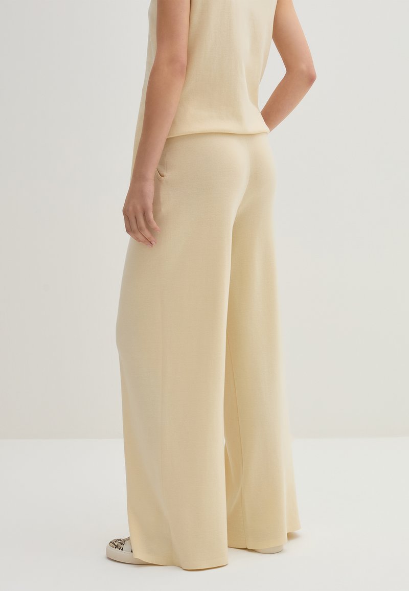 Pantaloni larghi beige realizzati in tessuto morbido, con una parte frontale liscia, tasche posteriori e una vestibilità rilassata. Abbinati a una maglia coordinata.