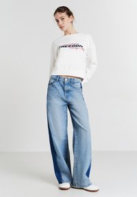 Felpa corta bianca con la scritta "FREEDOM" in nero e rosso. Jeans a gamba larga blu chiaro con pannelli laterali blu scuro. Sneakers bianche.