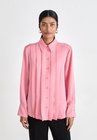 Lys rosa skjorte med krage, vertikale folder og lange ermer. Har små knapper og en jevn tekstur.