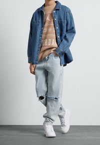 Camicia di jeans blu, indossata aperta sopra una felpa marrone chiaro con motivi di basket. Jeans azzurri slavati con orlo strappato. Scarpe da ginnastica bianche.