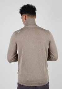Pull en coll roulé beige avec col et poignets côtelés, fabriqué en tissu doux. Présente un motif subtil et un petit logo sur l'épaule.