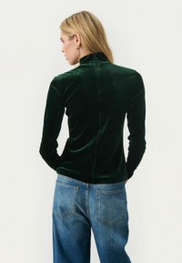 Grön sammet långärmad topp med hög krage; har en figurnära design och en söm längs mitten av ryggen. Matchas med blå denimjeans.