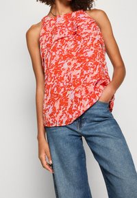Top senza maniche a fantasia rossa e rosa con un design a campana e un lieve dettaglio di rouches sul colletto, abbinato a jeans blu a gamba larga.