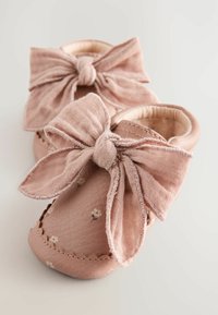 Rosa Leder-Babyschuhe mit einer großen Stoffschleife, floralen Mustern und gewellten Nähten entlang der Kanten. Weiche, strukturierte Oberfläche.