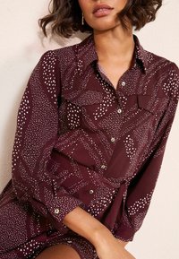 Bordo spalvos suknelė iš lengvos medžiagos su baltais taškeliais, turi apykaklinį iškirpimą, dvi krūtinės kišenes ir sagčių užsegimą.