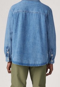 Jasnoniebieska koszula jeansowa z długimi rękawami, zapinanymi na guziki mankietami i prostym krojem. Posiada tylne karczek i jest noszona na oliwkowozielonych spodniach.