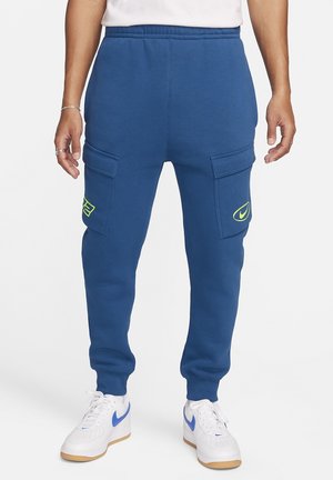 Osoba ubrana w niebieskie spodnie dresowe typu cargo z neonowymi logo na kieszeniach i białe sneakersy z niebieskimi znakami Nike, stojąca na białym tle.