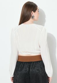 Weiße Bluse mit langen puffärmel und elastischem Saum, kombiniert mit einem schwarzen, strukturierten Rock mit Paisleymuster und braunem Bund.
